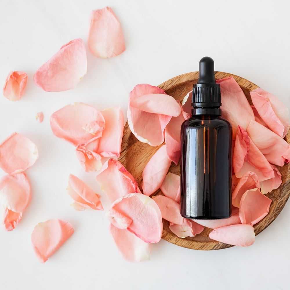 Essential oil dropper bottle with rose petals on wooden plate.