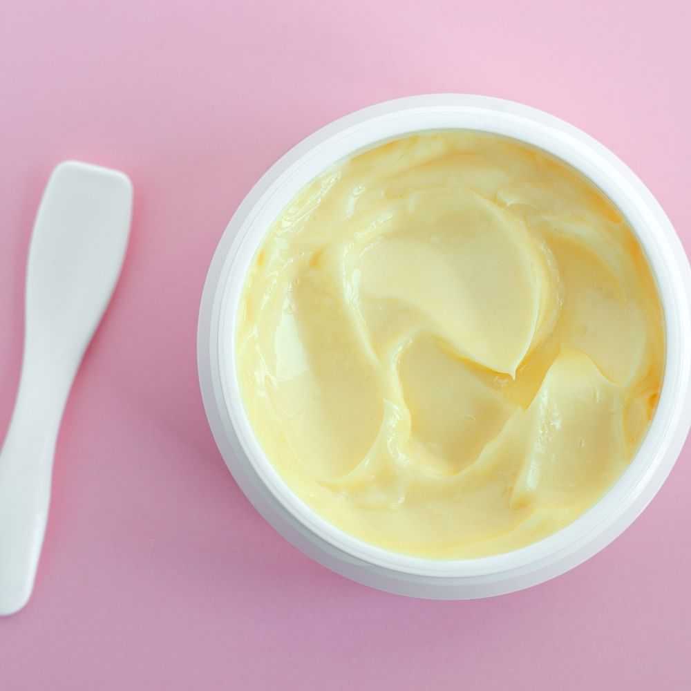 Open jar of cream with spatula on a pink background.